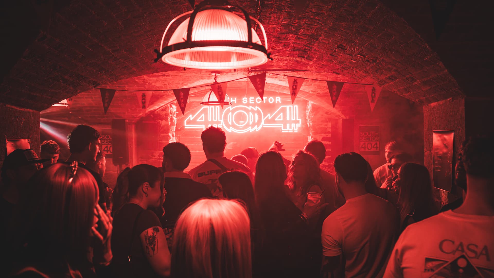 A crowd gathered under a red-lit 'South Sector 404' neon sign at a club night, photographed by Shore Break Media